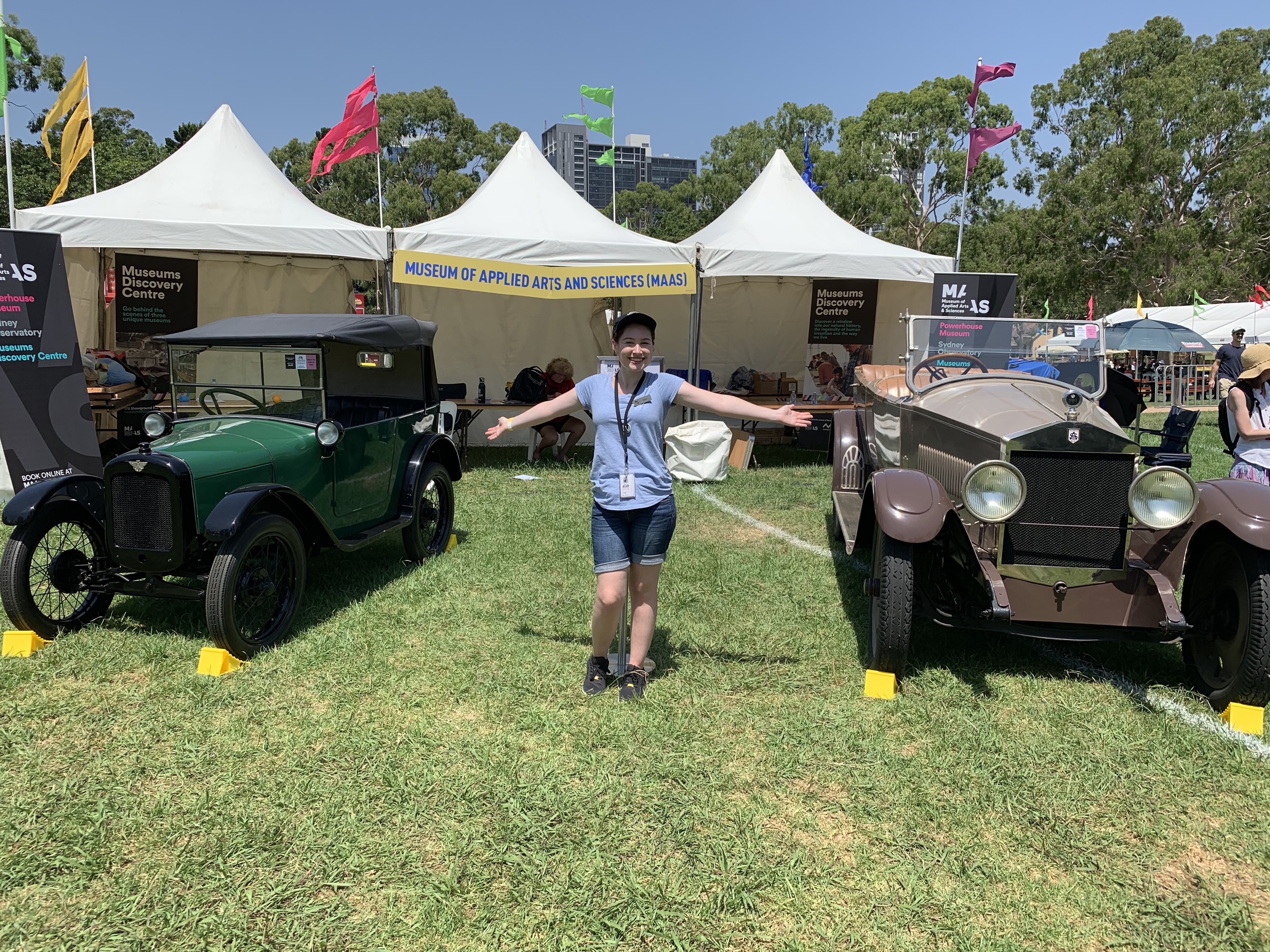 Small pale woman in a blue t-shirt, jean shorts and black cap stands on grass between two classic cars with her arms outstretched. Car on the left is green and black and the other on the right is light and dark brown. Behind her is a white fabric marquee with three roofed points and multicoloured flags blowing in the breeze. 
